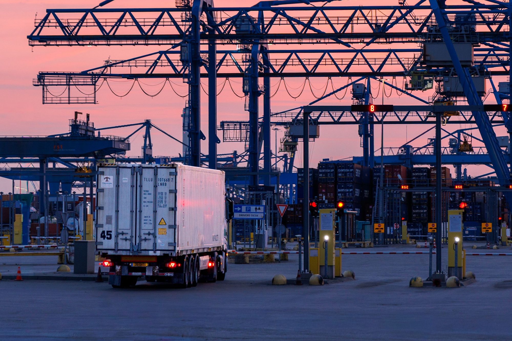 A container truck passing through a port terminal gate at sunset, gantry cranes behind
